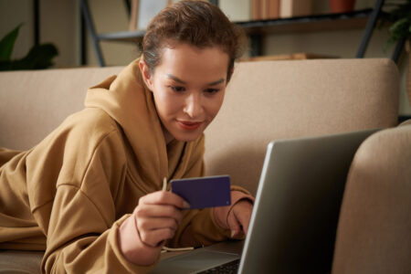 young woman laying on couch holding her credit card with her laptop open